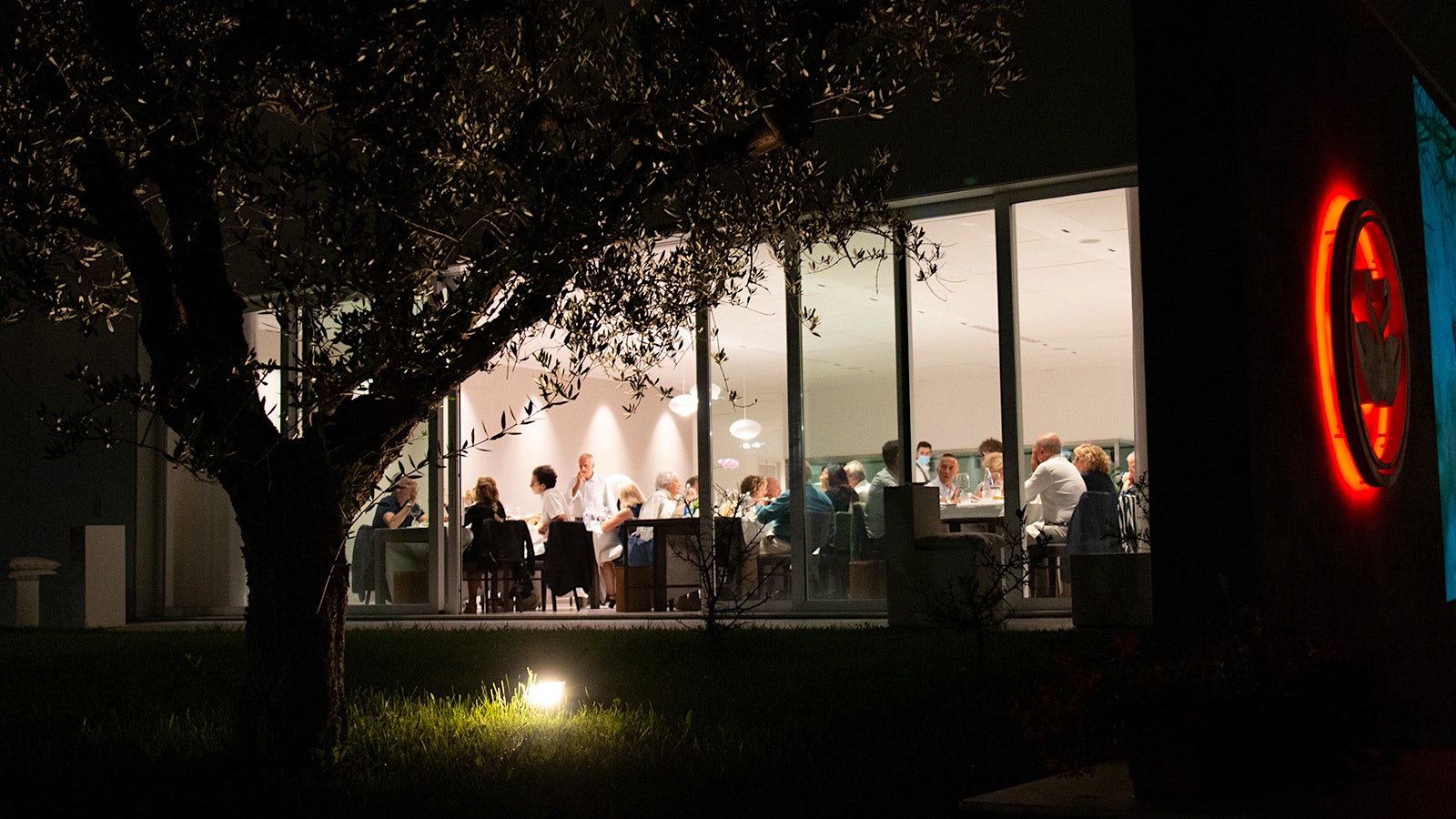 An exterior shot of people dining at Due Cigni Il Ristorante, with floor-to-ceiling windows and a white, modern-design interior
